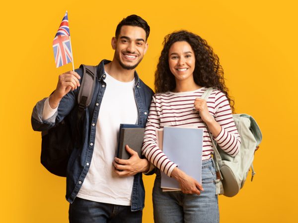 Study Abroad. Cheerful Young Arab Man And Woman With Backpacks Holding British Flag, Portrait Of Happy Smiling Students With Union Jack And Workbooks Over Yellow Studio Background, Copy Space